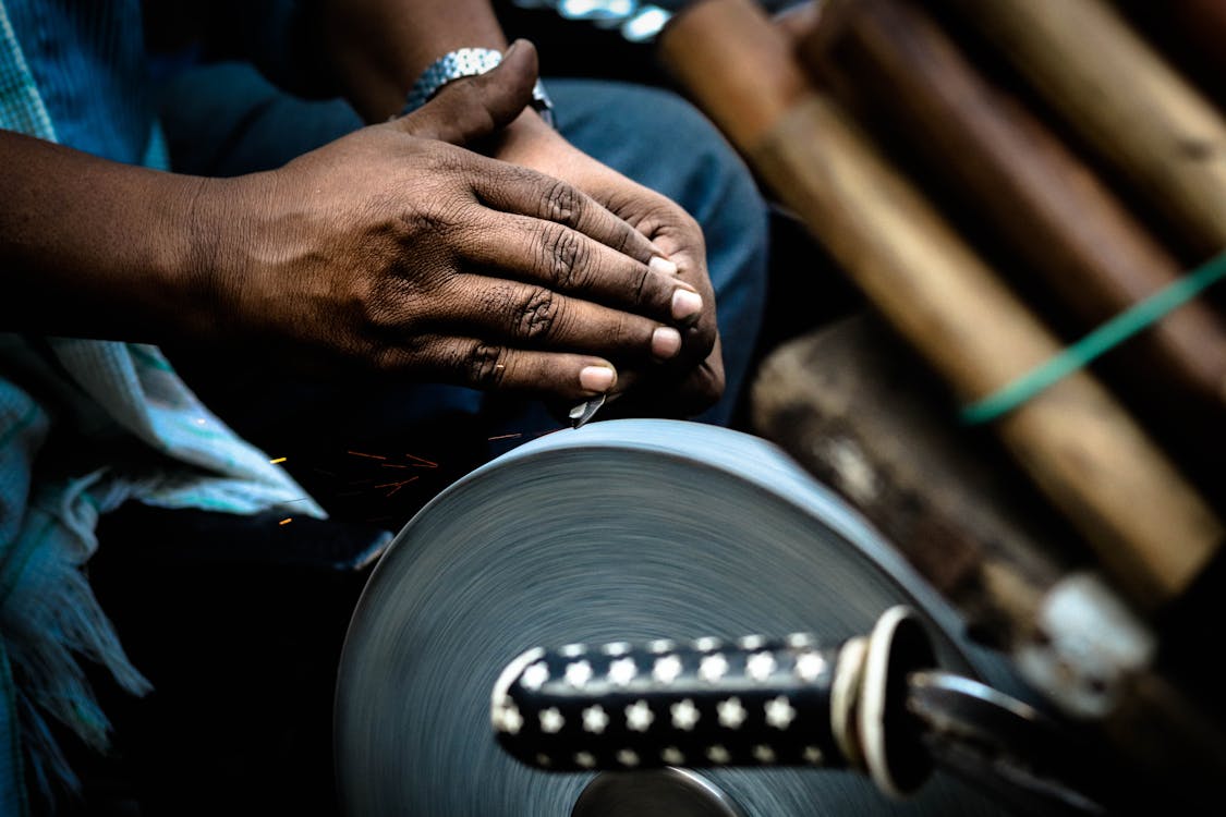 Main sujet: mains d’un artisan faisant tourner une meule sur un outil de coupe; étincelles visibles, ambianceatelier decoutellerie.