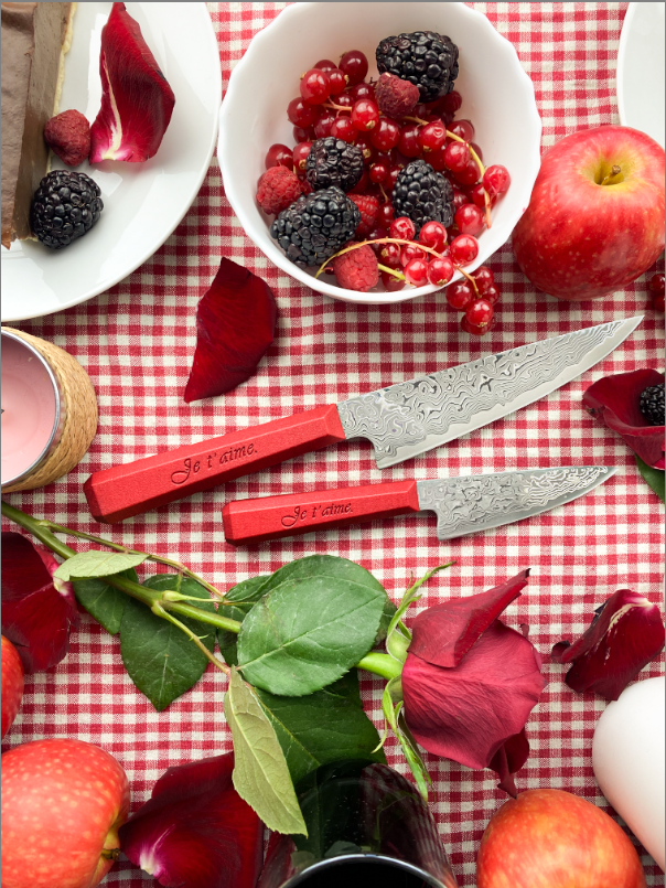 Couteaux damassés à lame argentée et manche rouge gravé « Je t’aime », posés sur une nappe à carreaux avec fruits rouges et pétales, bouquet de roses rouges.