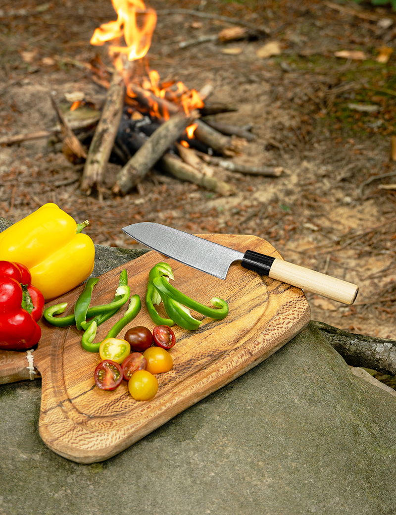 Couteau japonais sur planche en bois avec poivrons et tomates cerises, près d’un feu de camp en extérieur. fréquence entretien couteaux japonais intégrée.