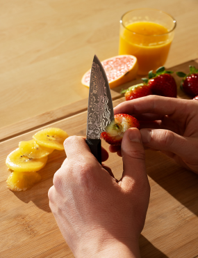 Personne aiguisant la lame d’un couteau sur planche en bois, tranches de fruits et jus d’orange en arrière-plan. Comment faire le tranchant d'un couteau.